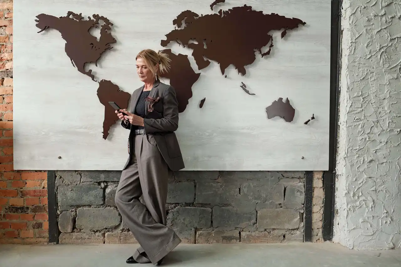 Serious mature female manager on international relationship standing against wall with map design and answering messages on phone